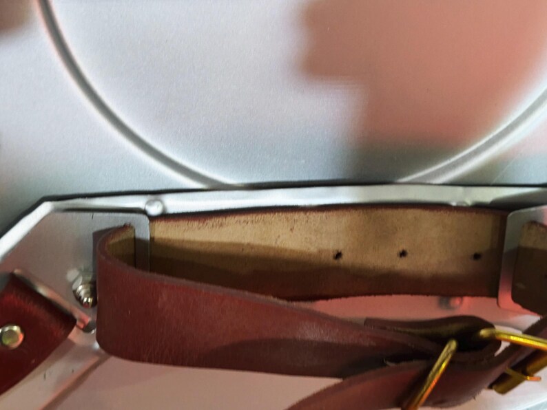 May include: Close-up of a brown leather strap with a gold-coloured buckle, attached to a white object with silver metal hardware. The leather strap has a natural edge and is secured with rivets.