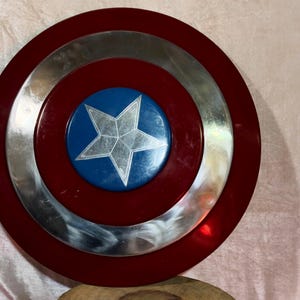 May include: A circular shield with a red border, a silver central ring, and a blue central disc. A silver star is in the middle. The shield is a prop or collectible.