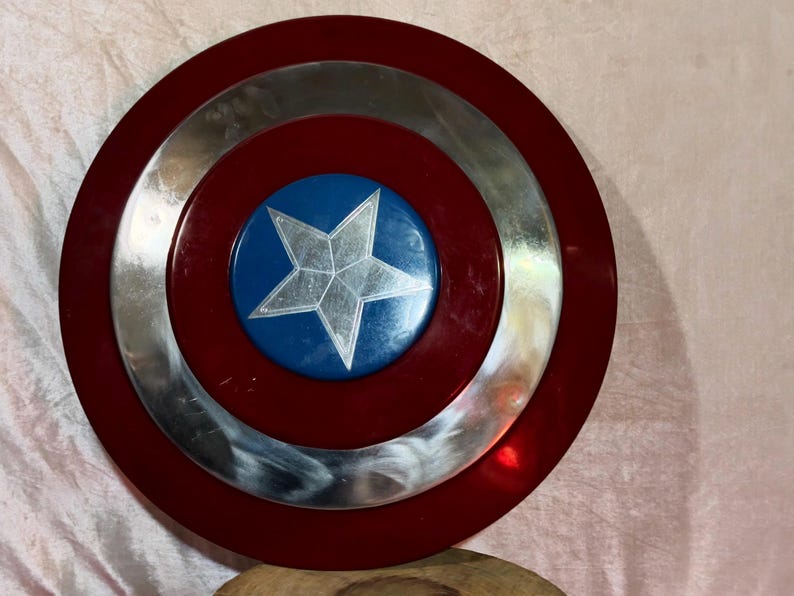 May include: A circular shield with a red border, a silver central ring, and a blue central disc. A silver star is in the middle. The shield is a prop or collectible.