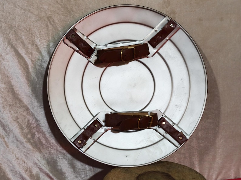May include: A silver shield featuring concentric circles and brown leather straps with gold buckles. The shield is circular and has a metallic finish. The diameter is approximately 60 cm.