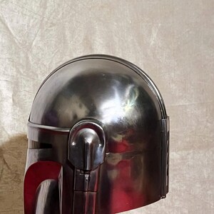 Może przedstawiać: Metalowy srebrny hełm z zaokrąglonym wierzchem i kanciastymi elementami. Hełm posiada wizjer i detale boczne, z czerwonym wnętrzem widocznym w obszarze wizjera. Prawdopodobnie element kostiumu lub przedmiot kolekcjonerski.