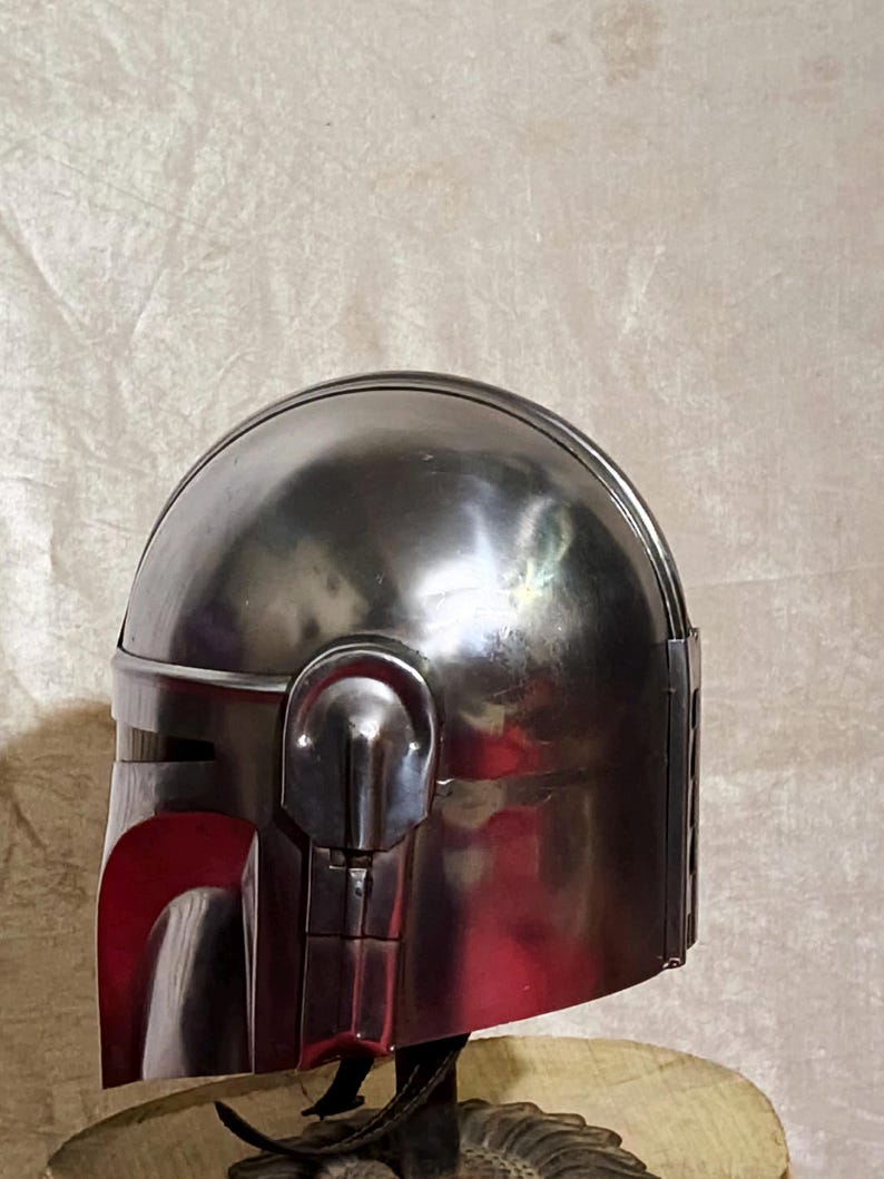 Może przedstawiać: Metalowy srebrny hełm z zaokrąglonym wierzchem i kanciastymi elementami. Hełm posiada wizjer i detale boczne, z czerwonym wnętrzem widocznym w obszarze wizjera. Prawdopodobnie element kostiumu lub przedmiot kolekcjonerski.