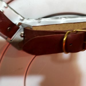 May include: Close-up of a brown leather strap with a gold-coloured buckle. The strap is attached to a silver metal frame. The leather has a textured appearance.