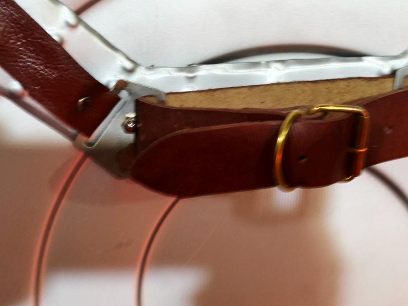 May include: Close-up of a brown leather strap with a gold-coloured buckle. The strap is attached to a silver metal frame. The leather has a textured appearance.