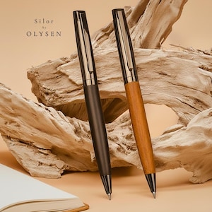 Könnte beinhalten: Zwei elegante Stifte, einer dunkelgrau und der andere holzfarben, werden vor einem Hintergrund aus Treibholz präsentiert. Die Stifte haben silberne Akzente und tragen die Aufschrift "Silor by Olysen". Ein Notizbuch ist unten links zu sehen.