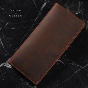 Pode incluir: Uma carteira de couro marrom com costuras visíveis e formato retangular. A carteira tem uma superfície lisa e uma borda marrom mais escura. O texto "Valten by OLYSEN" é visível no canto superior esquerdo.