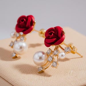 May include: Gold-coloured earrings with a red rose, a large white pearl, and small clear crystals. The earrings have a post-back closure. The earrings are set against a neutral background.