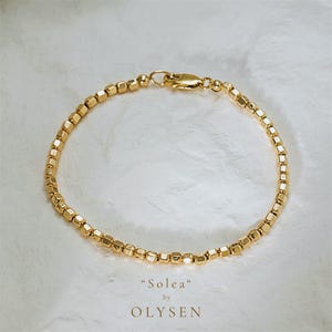Può includere: Un braccialetto dorato con una serie di piccole perline quadrate. Il braccialetto è chiuso con una chiusura. Il testo "Solea" by OLYSEN è visualizzato sotto.