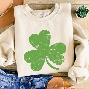 May include: Cream-colored sweatshirt featuring a large, distressed green shamrock design. The long-sleeved top is laid on a wooden surface with blue denim jeans, a brown hat, and sandals.