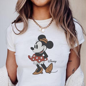 May include: White crop top featuring a graphic of Minnie Mouse in a red polka dot skirt and brown shoes. The Disney logo is printed on the shirt. The shirt has short sleeves and a crew neck.