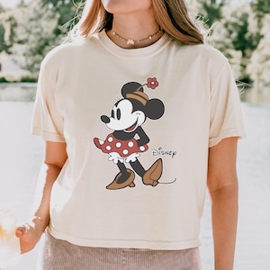 May include: Ivory-colored t-shirt featuring a Minnie Mouse graphic. The cartoon character is depicted in a red polka-dot dress, brown shoes, and a matching hat. The word "Disney" is printed below the character. The shirt has a Comfort Colors label.