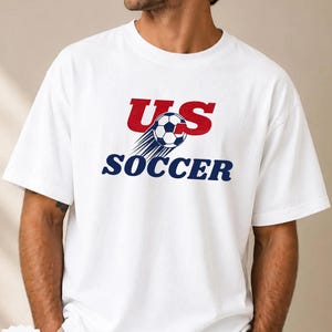 Op de afbeelding: Wit T-shirt met de tekst "US SOCCER" in rood en marineblauw, en een voetbalafbeelding. Het shirt heeft een ronde hals.