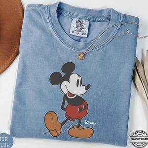 May include: Pale blue t-shirt with a Mickey Mouse graphic. The cartoon character is depicted in his classic pose. The shirt has a crew neck and short sleeves. The text "Disney" is printed below the character. The shirt is labeled "Comfort Colors" and made of 100% cotton.
