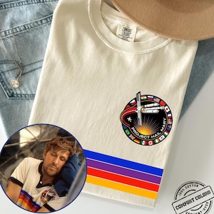 May include: Cream-colored t-shirt with a circular graphic featuring a rocket and the text "PROJECT HAIL MARY." Below the graphic are horizontal stripes in blue, purple, red, and yellow. The shirt is made of 100% cotton.