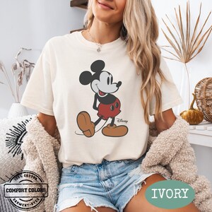 May include: Ivory t-shirt with a Mickey Mouse graphic in black, red, and brown. The shirt is labelled "Comfort Colors" and "Garment-Dyed". The colour "Ivory" is indicated. The t-shirt is a casual wear item.