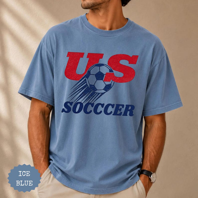 K&ouml;nnte beinhalten: Eisblaues T-Shirt mit rotem "US"-Schriftzug und einem Fu&szlig;ball-Design. Das Wort "SOCCER" ist darunter in Marineblau gedruckt. Das Shirt hat eine lockere Passform.