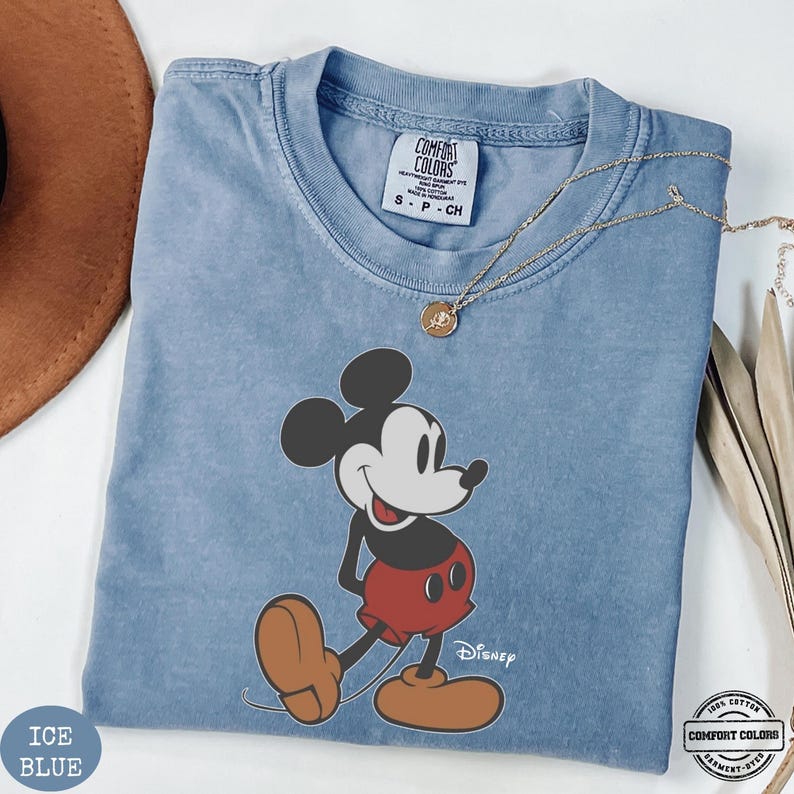 May include: Pale blue t-shirt with a Mickey Mouse graphic. The cartoon character is depicted in his classic pose. The shirt has a crew neck and short sleeves. The text "Disney" is printed below the character. The shirt is labeled "Comfort Colors" and made of 100% cotton.