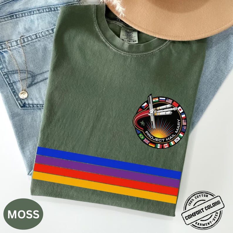 May include: Moss green t-shirt with a colorful horizontal stripe design and a circular patch that reads "Project Hail Mary." The patch features a spaceship and flags. The shirt is made of 100% cotton and is garment-dyed.