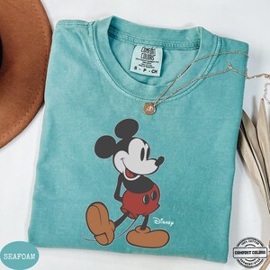 May include: Seafoam green t-shirt with a large Mickey Mouse graphic. The cartoon character is in a classic pose. The shirt has a crew neck and short sleeves. The Disney logo is printed below the character. A gold necklace with a pendant is draped across the shirt.