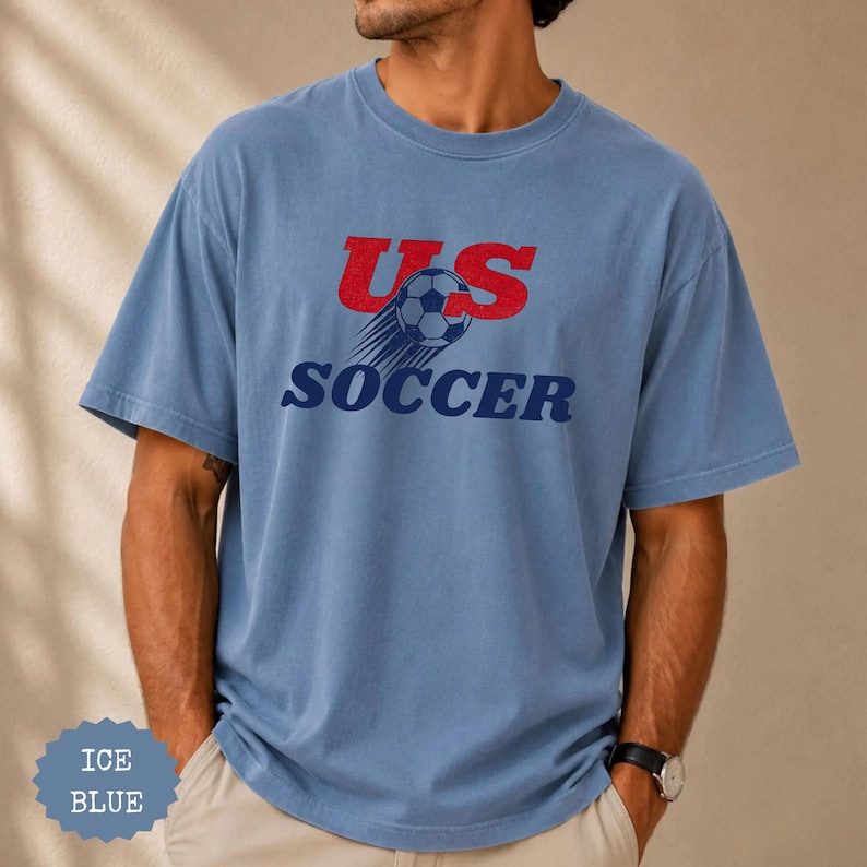 Op de afbeelding: Ijsblauwe t-shirt met de tekst "US Soccer" in rood en marineblauw. Het ontwerp bevat een voetbal met bewegingslijnen. Het shirt is een casual stijl met korte mouwen.