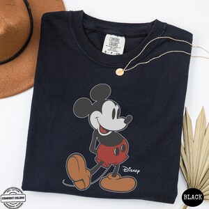 May include: A navy blue t-shirt with a graphic of Mickey Mouse. The cartoon character is depicted in his iconic pose. The word "Disney" is printed below the character. A gold necklace and a brown hat are also visible.