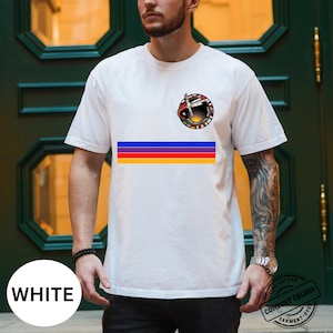 May include: White t-shirt featuring a colorful horizontal stripe design and a circular graphic with text and a rocket ship. The shirt also has a small graphic on the upper left chest. The shirt is made of a soft, comfortable material.
