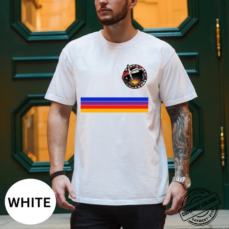May include: White t-shirt featuring a colorful horizontal stripe design and a circular graphic with text and a rocket ship. The shirt also has a small graphic on the upper left chest. The shirt is made of a soft, comfortable material.