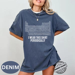 Może przedstawiać: Niebieska koszulka dżinsowa z białym wzorem układu okresowego i napisem "I wear this shirt periodically." Koszulka jest wykonana w 100% z bawełny i farbowana w kawałku.