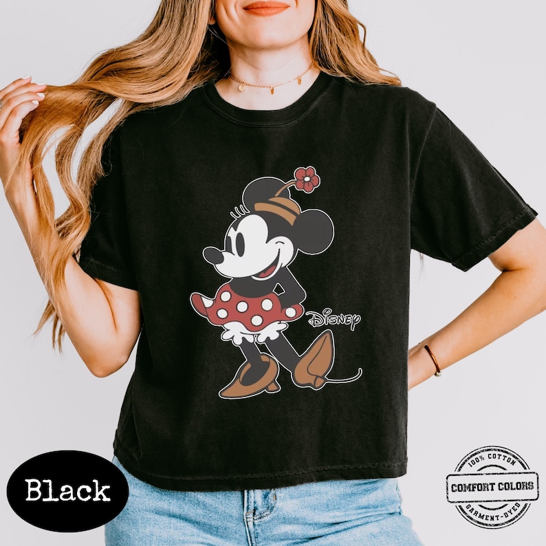 May include: Black t-shirt featuring a Minnie Mouse graphic. The cartoon character is depicted in a red and white polka-dot dress, brown shoes, and a hat with a flower. The word "Disney" is printed below the graphic. The shirt also has the words "100% Cotton" and "Comfort Colors Garment-Dyed".