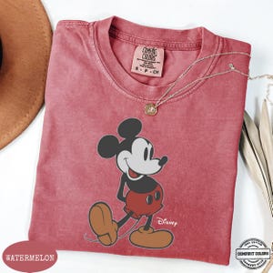 May include: A watermelon-coloured t-shirt featuring a graphic of Mickey Mouse. The shirt has a crew neck and short sleeves. The Disney logo is visible. A gold necklace is draped across the shirt. The shirt is labelled "Comfort Colors".