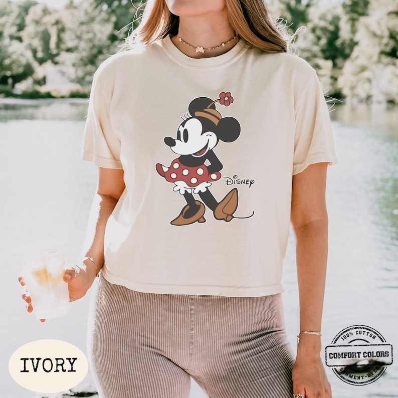 May include: Ivory-colored t-shirt featuring a Minnie Mouse graphic. The cartoon character is depicted in a red polka-dot dress, brown shoes, and a matching hat. The word "Disney" is printed below the character. The shirt has a Comfort Colors label.