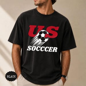 K&ouml;nnte beinhalten: Schwarzes T-Shirt mit dem Text "US SOCCER" in Rot und Wei&szlig;. Das Design beinhaltet einen Fu&szlig;ball mit Bewegungslinien. Das Shirt ist dunkel und hat einen Vintage-Look.