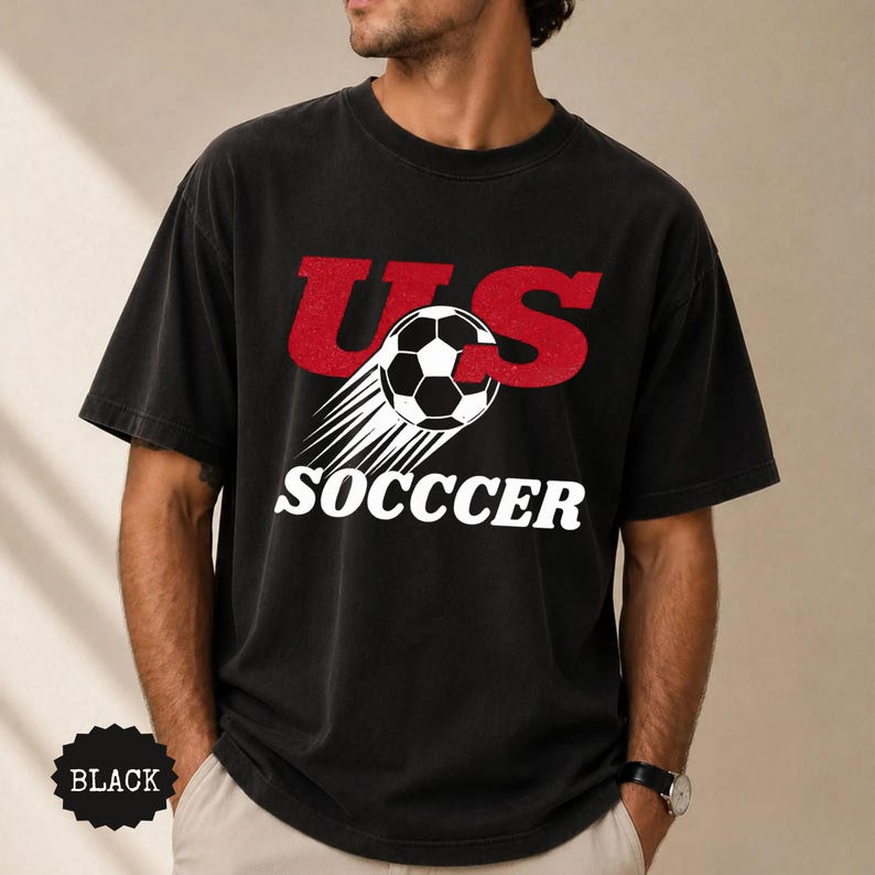 K&ouml;nnte beinhalten: Schwarzes T-Shirt mit dem Text "US SOCCER" in Rot und Wei&szlig;. Das Design beinhaltet einen Fu&szlig;ball mit Bewegungslinien. Das Shirt ist dunkel und hat einen Vintage-Look.