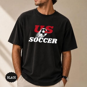 Op de afbeelding: Zwart T-shirt met de tekst "US SOCCER" in wit en rood, met een voetbal met bewegingslijnen. Het shirt heeft een vintage uitstraling en is donker van kleur.