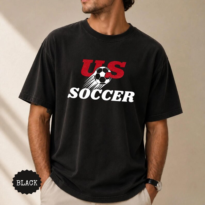 Op de afbeelding: Zwart T-shirt met de tekst "US SOCCER" in wit en rood, met een voetbal met bewegingslijnen. Het shirt heeft een vintage uitstraling en is donker van kleur.