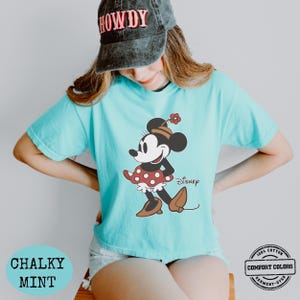 May include: A light blue t-shirt featuring a Minnie Mouse graphic. The cartoon character is wearing a red polka dot skirt, brown shoes, and a hat with a flower. The shirt has the word "Disney" printed on it. The shirt is labeled "Chalky Mint" and "Comfort Colors".