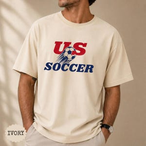 Op de afbeelding: Ivoor kleurig T-shirt met de tekst "US SOCCER" in rood en blauw. Het ontwerp bevat een voetbal met bewegingslijnen. Het shirt is een casual stijl.