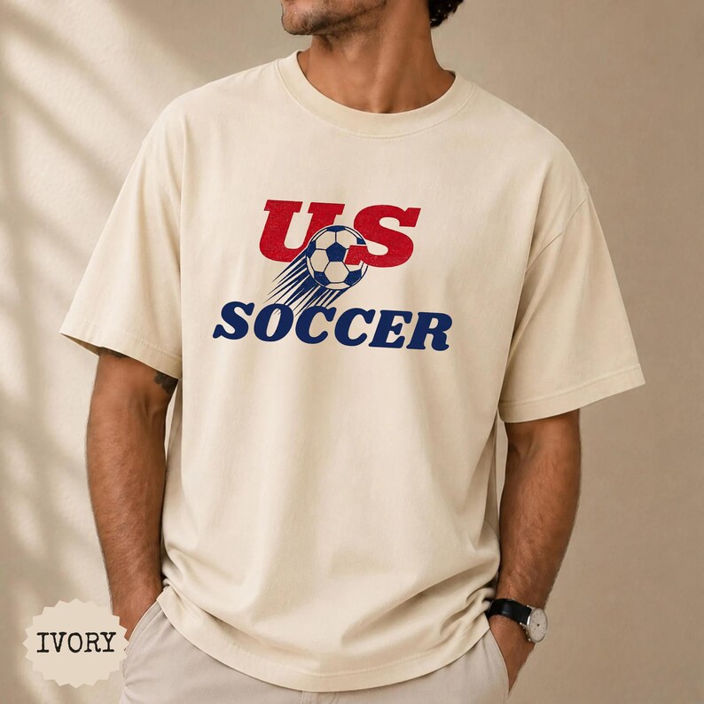 Op de afbeelding: Ivoor kleurig T-shirt met de tekst "US SOCCER" in rood en blauw. Het ontwerp bevat een voetbal met bewegingslijnen. Het shirt is een casual stijl.