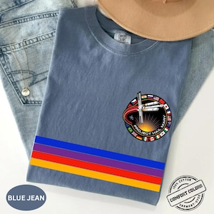 May include: Blue jean colored t-shirt with a colorful horizontal stripe design and a circular graphic that reads "Project Hail Mary". The shirt is made of 100% cotton and is garment-dyed.