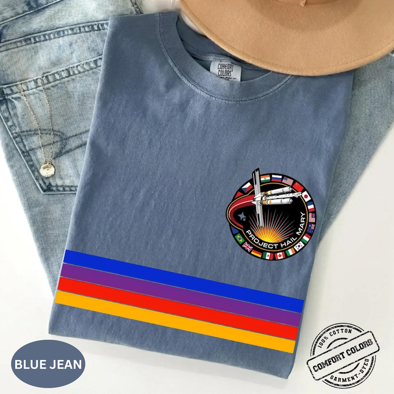 May include: Blue jean colored t-shirt with a colorful horizontal stripe design and a circular graphic that reads "Project Hail Mary". The shirt is made of 100% cotton and is garment-dyed.
