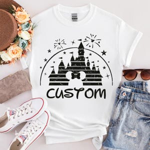 May include: White t-shirt with a black graphic of a castle, fireworks, stars, and a Mickey Mouse bow. The word "CUSTOM" is printed below the castle design. The shirt is displayed with sunglasses, denim shorts, and white sneakers.