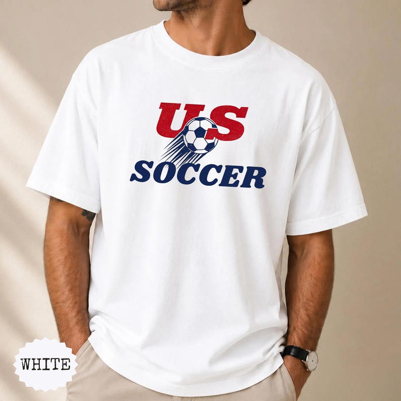 Op de afbeelding: Wit T-shirt met de tekst "US SOCCER" in rood en marineblauw, en een voetbalafbeelding. Het shirt heeft een ronde hals.