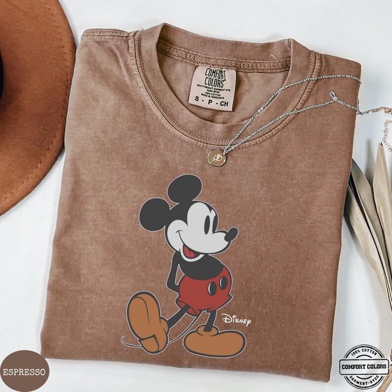 May include: Espresso-coloured t-shirt featuring a graphic of Mickey Mouse in his classic pose. The shirt has a crew neck and short sleeves. A silver necklace with a small pendant is draped across the shirt. The shirt is labelled "Comfort Colors" and "100% Cotton".