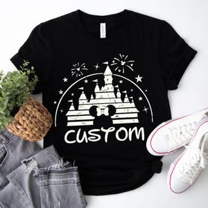 May include: Black t-shirt with a white graphic featuring a castle, fireworks, and Mickey Mouse ears. The word "custom" is printed below the graphic. A pair of white sneakers and gray jeans are next to the shirt.