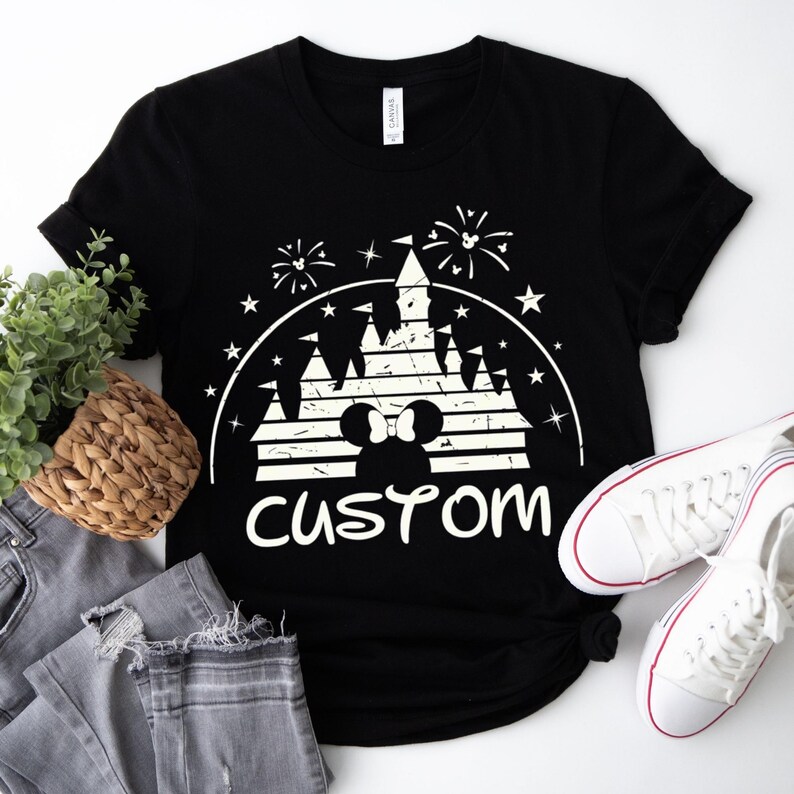 May include: Black t-shirt with a white graphic featuring a castle, fireworks, and Mickey Mouse ears. The word "custom" is printed below the graphic. A pair of white sneakers and gray jeans are next to the shirt.