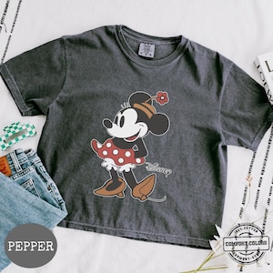 May include: A dark gray t-shirt featuring a graphic of Minnie Mouse in a red polka dot dress and brown shoes. The word "Disney" is printed below the character. The shirt is made of 100% cotton and is garment-dyed.