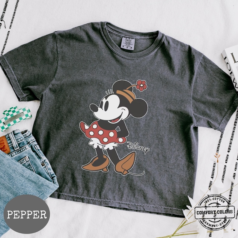 May include: A dark gray t-shirt featuring a graphic of Minnie Mouse in a red polka dot dress and brown shoes. The word "Disney" is printed below the character. The shirt is made of 100% cotton and is garment-dyed.