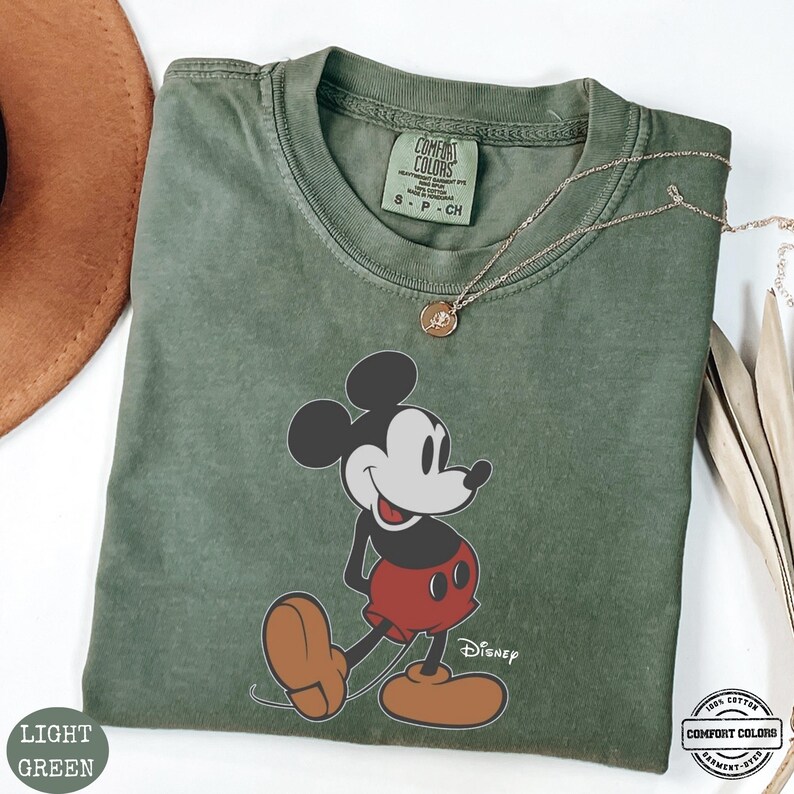 May include: Light green t-shirt with a Mickey Mouse graphic in red shorts and brown shoes. The shirt has the text "Disney" and "Comfort Colors" printed on it. A gold necklace is draped across the shirt.