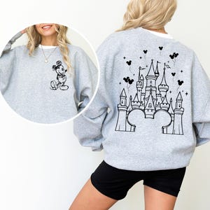 Puede incluir: Una sudadera gris claro con un dise&ntilde;o de Mickey Mouse en blanco y negro en la parte delantera. La parte trasera de la sudadera muestra un dise&ntilde;o de castillo en blanco y negro, orejas de Mickey Mouse y globos.
