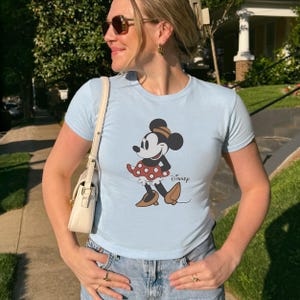 May include: Light blue t-shirt featuring a Minnie Mouse graphic. The cartoon character is depicted in a red and white polka dot dress, brown shoes, and a matching hat. The word "Disney" is printed below the graphic.
